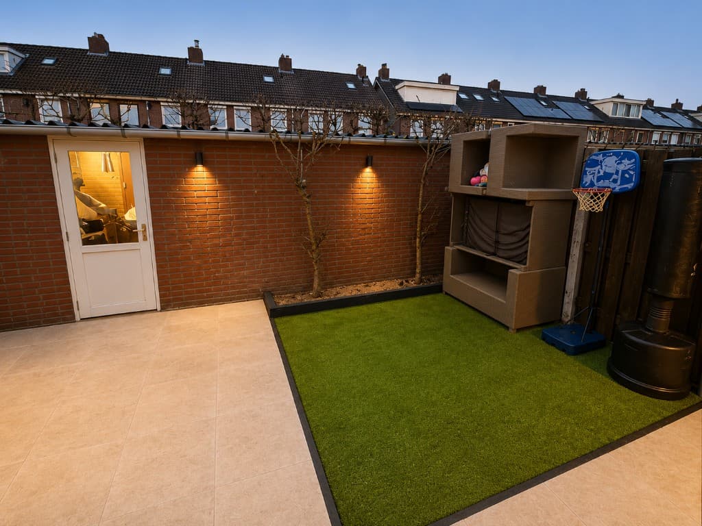 Vernieuwde tuin en terras bij een woning in Nederland, met bestrating, kunstgras en buitenverlichting bij schemer.