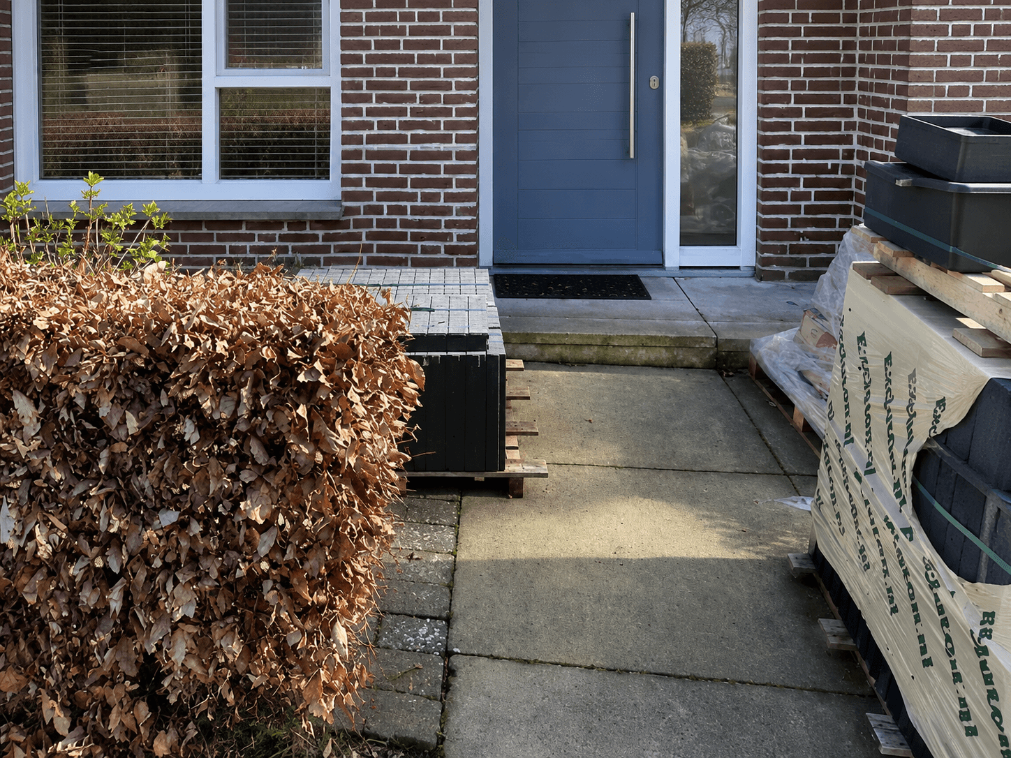 Front garden before the works, with dark slabs, a dried-out hedge and stored materials.